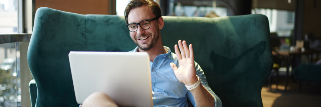 Man wearing glasses seated on a teal sofa with a laptop, using Biteable video maker, waving at the camera.