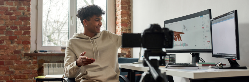 A young man records a video in a brick-walled office, gesturing towards graphs on his computer screen, with a Biteable video maker camera in the foreground.